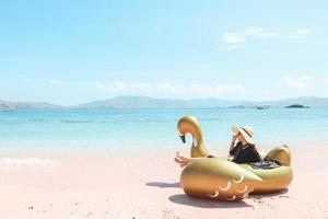 uma garota de chapéu de verão sentado no cisne inflável enquanto aprecia a vista para o mar na praia de areia rosa foto