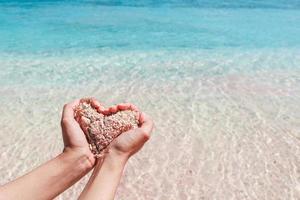 mão fazendo formato de coração de areia rosa com água transparente na praia foto