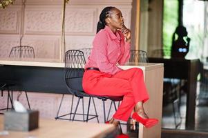 bonito tranças negócio afro-americano senhora brilhante mandão pessoa amigável desgaste escritório vermelho camisa e calças, sentado na cadeira. foto