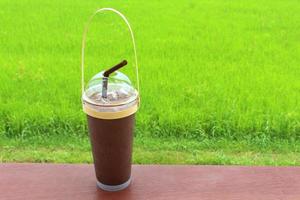 bebida de chocolate escuro legal em copo de plástico transparente com palha marrom e alça de bambu na mesa de madeira com fundo de campo de arroz. foto