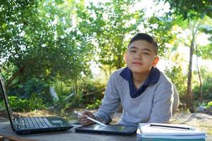 menino olhando para a câmera estudando online em uma mesa de madeira com computador portátil e tablet de manhã em uma casa rural. conceito área rural de educação on-line e trabalho em casa foto