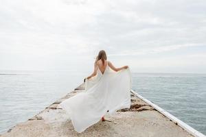 uma jovem linda em um vestido longo cor de leite caminha ao longo da praia e cais no contexto do mar. foto