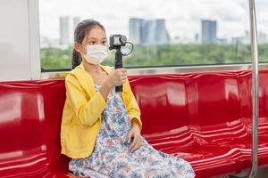 menina segurando câmera de ação, criança usa máscara facial durante surto de coronavírus e gripe, viajando de trem. foto