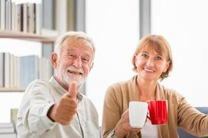 casal sênior dentro de casa durante uma pausa para o café, sorrindo casal de idosos mostrando os polegares segurando xícaras de café foto