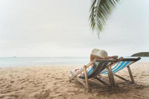 jovem mulher da ásia relaxando na cadeira de praia braço para cima a mão com chapéu flexível foto
