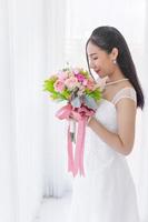 uma noiva asiática vestida com um vestido de noiva branco fica sorrindo brilhantemente em uma mão segurando um lindo buquê de flores. foto