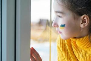 uma criança triste olha para a janela com a bandeira da ucrânia pintada na bochecha, preocupações e medo. ajuda humanitária às crianças, paz mundial, segurança. foto