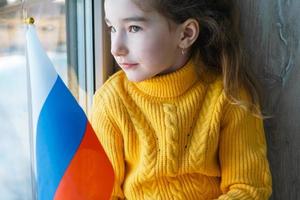 uma criança triste na janela com a bandeira da rússia, se preocupa com lágrimas nos olhos. conflito entre rússia e ucrânia, medo foto