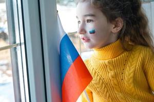 uma criança triste na janela com a bandeira da rússia, se preocupa com lágrimas nos olhos. conflito entre rússia e ucrânia, medo foto