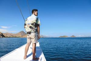 homem asiático em óculos de sol em pé no barco enquanto olha para o mar com paisagem marinha e colinas em labuan bajo foto