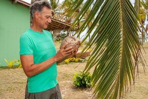 homem sênior sob um coqueiro segurando um coco que caiu da árvore foto