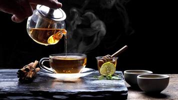 derramando chá de ervas aromáticas quente do bule em xícara de chá de vidro com vapor e várias ervas na placa de pedra preta com piso de mesa de madeira em fundo escuro, foco seletivo