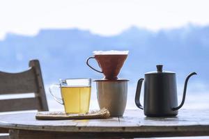 xícara de vidro de chá aromático quente com xícara de café de gotejamento em mesa redonda de madeira contra fundo de vista ribeirinha na hora da manhã foto
