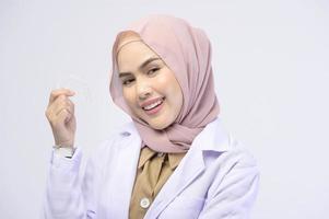 dentista feminino muçulmano segurando aparelho invisalign sobre estúdio de fundo branco, saúde bucal e conceito ortodôntico. foto