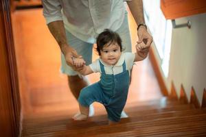 criança bonitinha conceito de família criança, bebê aprendendo a andar com pai e mãe para ajudar a cuidar e segurar a mão, primeiro passo com apoio dos pais na infância, pequeno retrato de amor menino em casa foto