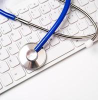 estetoscópio azul no teclado do computador no fundo da mesa branca. conceito de tecnologia de tratamento de informações médicas on-line, close-up, macro, copie o espaço foto