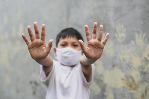 menino asiático com máscara médica mostrando parada de gesto com a mão com impressão de mão no fundo da parede de cimento foto