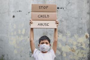 menino asiático em máscara médica levantando quadro de cartas diz parar a campanha de abuso infantil com impressão manual no fundo da parede de cimento