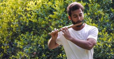 músico indiano masculino tocando bansuri vestindo camiseta branca. tiro ao ar livre em fundo verde foto