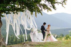 a noiva e o noivo estão abraçando sob um velho carvalho. sessão de fotos de casamento nas montanhas. ao lado deles está preparada a decoração para a cerimônia.