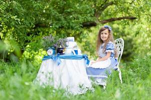 uma menina bonita na fantasia alice do país das maravilhas realiza uma festa de chá em sua mesa mágica. fotografado na natureza. foto