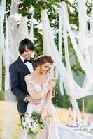 a noiva e o noivo estão abraçando sob um velho carvalho. sessão de fotos de casamento nas montanhas. ao lado deles está preparada a decoração para a cerimônia.