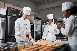 equipe gourmet profissional, três jovens chefs em uniformes e aventais de cozinheiro branco amassam massa de pastel e ovos, preparam pão e alimentos frescos de padaria, assando no forno na cozinha do restaurante de aço inoxidável. foto