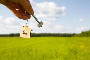 pingente de madeira de uma casa e chave. fundo do céu e campo. sonho de casa, construção de uma casa de campo no campo, plano e projeto, entrega do projeto, mudança para uma casa nova. espaço de cópia foto