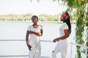 duas garotas afro-americanas elegantes e modernas, usam roupas brancas contra o lago. moda de rua de jovens negros. foto
