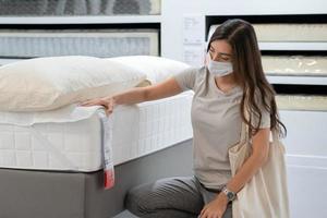 mulher usando máscara protetora selecionando colchão de cama para mudar de casa ou decorar novo quarto. novas compras normais durante a pandemia de coronavírus. foto