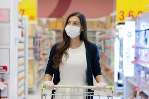 mulher usando máscara facial médica e luva de borracha empurra o carrinho de compras na loja de departamentos do supermercado. garota olhando para prateleira de supermercado. novo estilo de vida normal durante o coronavírus foto