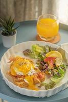 close-up em um café da manhã ou brunch, ovos benedict servem com bacon frito e torradas e salada de legumes para acompanhamento em um prato branco ou prato em pano branco com faixa verde e vem com suco de laranja foto