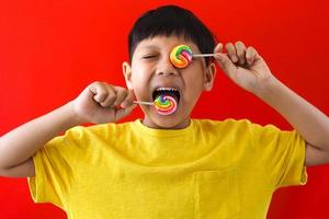 lindo menino asiático lambendo e segurando doces de pirulito em fundo vermelho foto