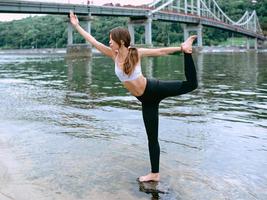 jovem apta em roupas esportivas em diferentes asanas de ioga ao ar livre na praia à beira do rio. conceito de ioga, natureza, cidade e esporte foto