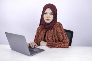 jovem mulher islâmica asiática está sentada trabalhando no laptop com olhar sério sobre o fundo branco isolado da câmera. foto