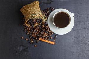grãos de café em saco marrom com xícara de café em fundo preto. foto