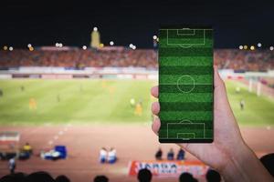 mão segurando o smartphone com fundo de estádio de campo de futebol desfocado. foto