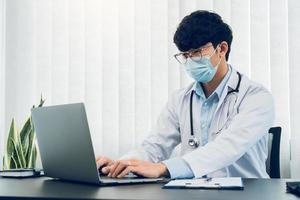 médico estava sentado no escritório trabalhando em um laptop enquanto usava uma máscara. foto