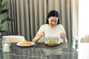 jovem asiática sorrindo enquanto ela pega uma salada em um prato e come alegremente em casa. foto