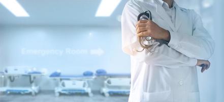 médico de medicina com estetoscópio na mão, confiante em frente à sala de emergência, leitos de pacientes atrás dele, fundo hospitalar foto