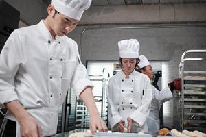 equipe gourmet profissional, três jovens chefs em uniformes e aventais de cozinheiro branco amassam massa de pastel e ovos, preparam pão e alimentos frescos de padaria, assando no forno na cozinha do restaurante de aço inoxidável. foto