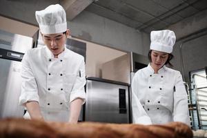 dois chefs asiáticos profissionais em uniformes e aventais de cozinheiro branco amassam massa e ovos, preparam pão, biscoitos e alimentos frescos de padaria, assando em um forno em uma cozinha de aço inoxidável de restaurante. foto