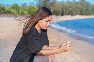 linda garota adolescente asiática pensando sozinha e assistindo a concha branca sentada na areia da praia foto