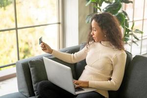 mulher latina usando laptop e segurando um cartão de crédito para fazer compras no sofá foto