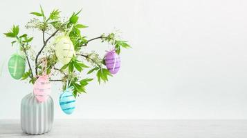 um buquê de galhos verdes com folhas frescas florescendo em um vaso em cima da mesa é decorado com ovos de páscoa coloridos. decoração no interior da casa para a páscoa. espaço de cópia