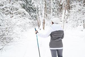 esquiador uma mulher em uma jaqueta de membrana com bastões de esqui nas mãos com as costas no contexto de uma floresta de neve. esqui cross-country na floresta de inverno, esportes ao ar livre, estilo de vida saudável. foto