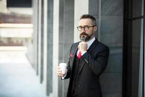empresário hipster em pé de terno com uma xícara de café perto do escritório ao ar livre foto