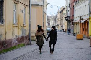 um jovem casal da moda caminha na cidade, a jovem tem sacolas de compras no braço e o homem uma xícara de café na mão foto