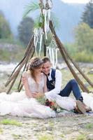 um atraente casal recém-casado, um momento feliz e alegre. um homem e uma mulher se barbeiam e se beijam com roupas de férias. cerimônia de casamento de estilo boêmio na floresta ao ar livre.