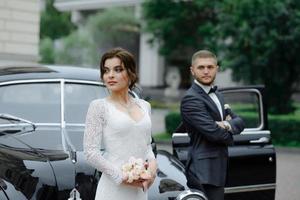 elegante linda noiva e belo noivo abraçando no elegante carro preto na luz. vista incomum de volta. casal de noivos de luxo em estilo retrô. foto